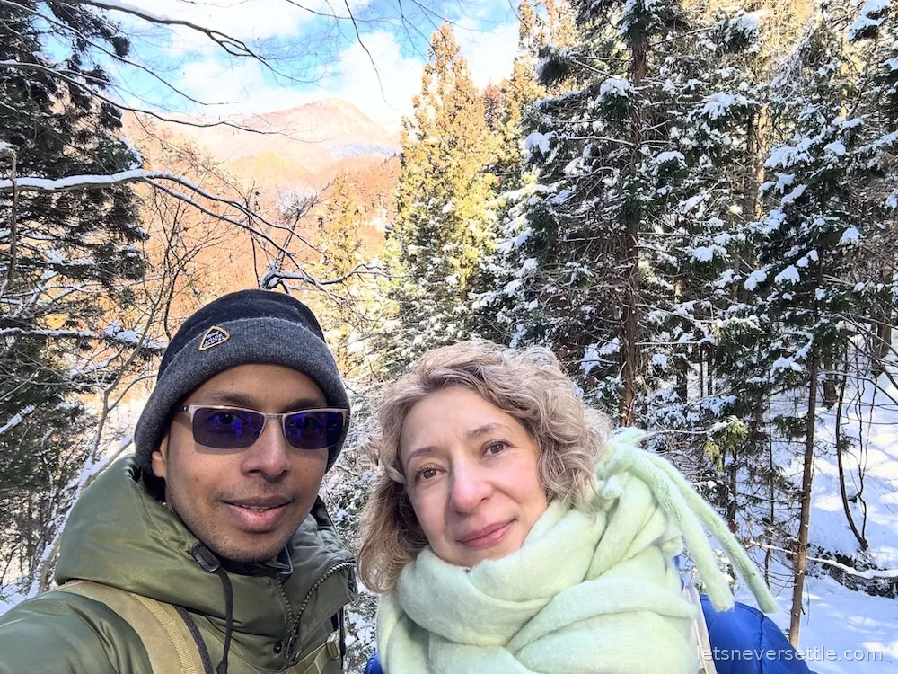 Selfie of Dileep in a bobble hat and sunglasses and Inga in a pale green scarf, snowy pines and a mountain ridge behind them