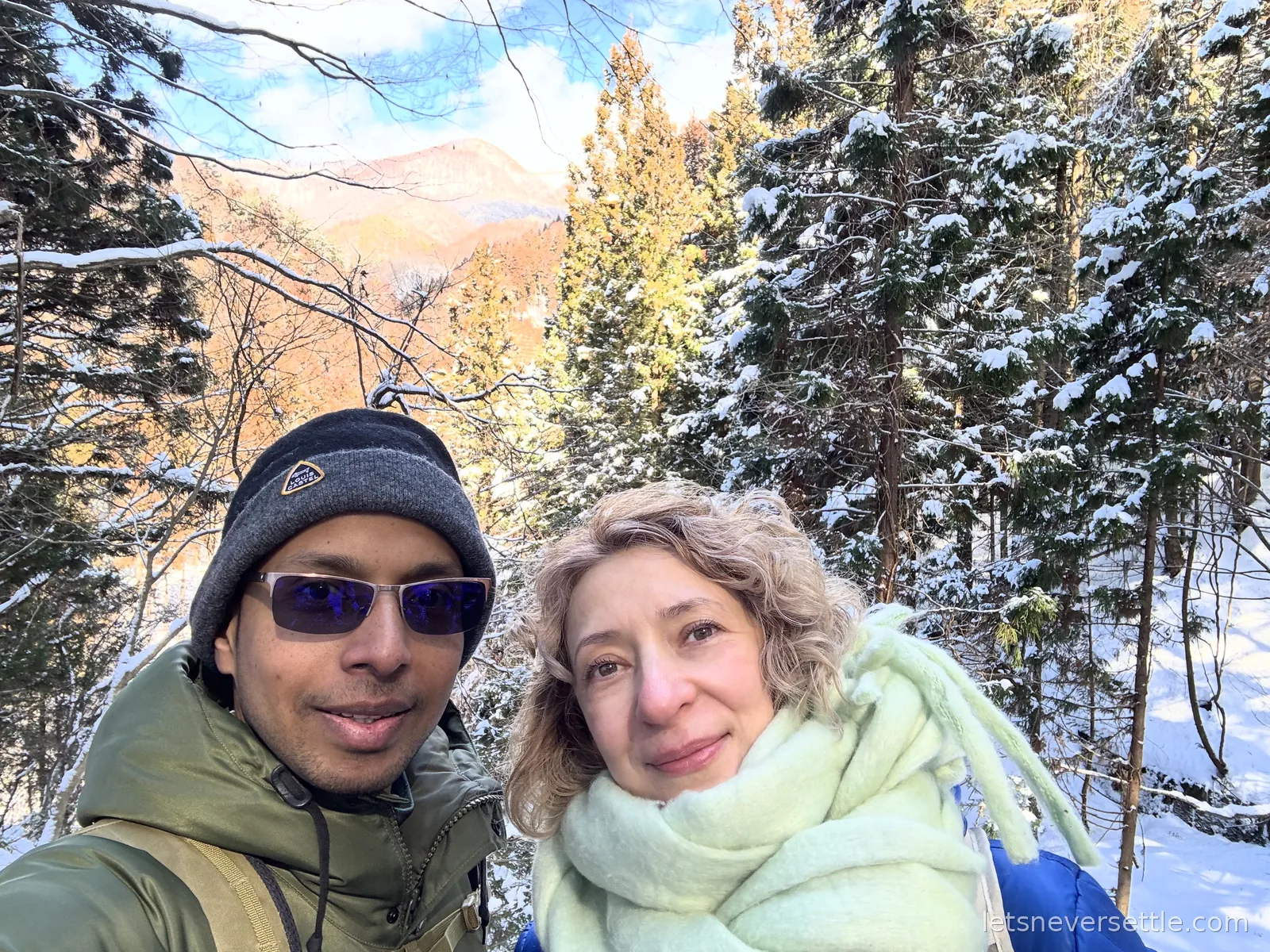 Selfie of Dileep in a bobble hat and Inga in a pale green scarf on a snowy forest trail with sunlit mountains behind
