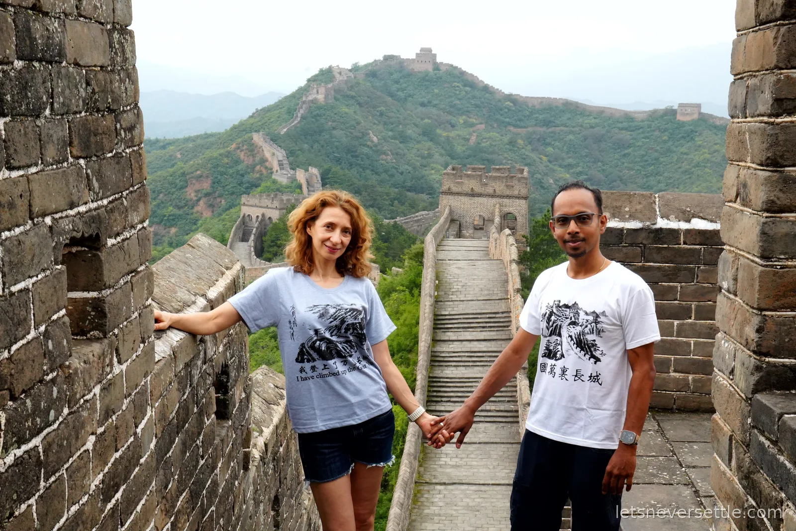 Inga and Dileep holding hands in a ruined watchtower doorway, the wall and its stairs stretching over the hills behind them