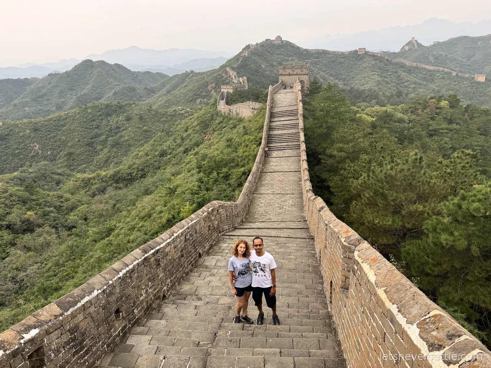 View down a long straight stretch of the Jinshanling wall walkway, steep stairs rising to a brick watchtower ahead