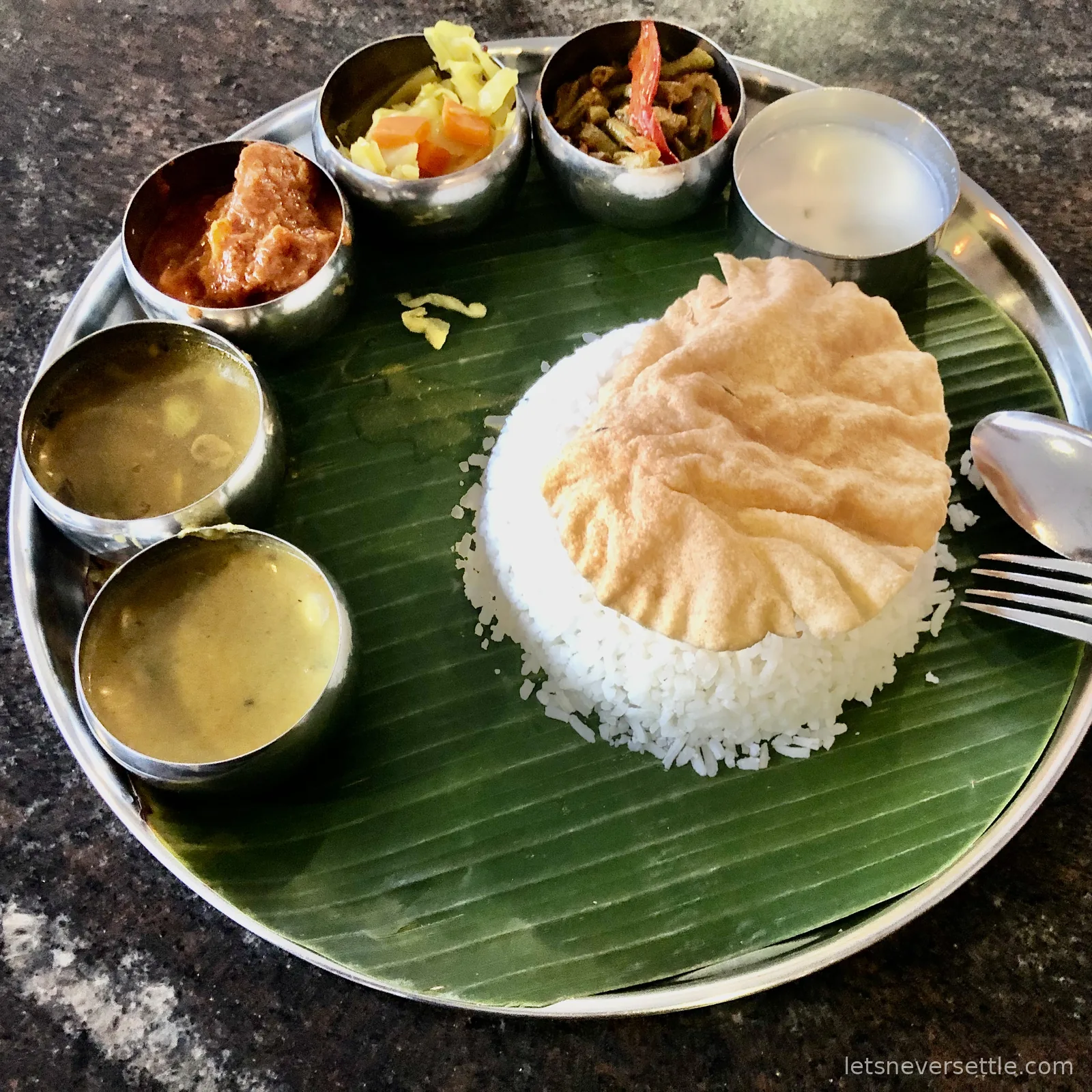 A banana leaf rice meal in Johor Bahru: white rice and a papadum on a banana leaf in a steel tray, ringed by small steel pots of curry, dhal, cabbage, okra and rasam