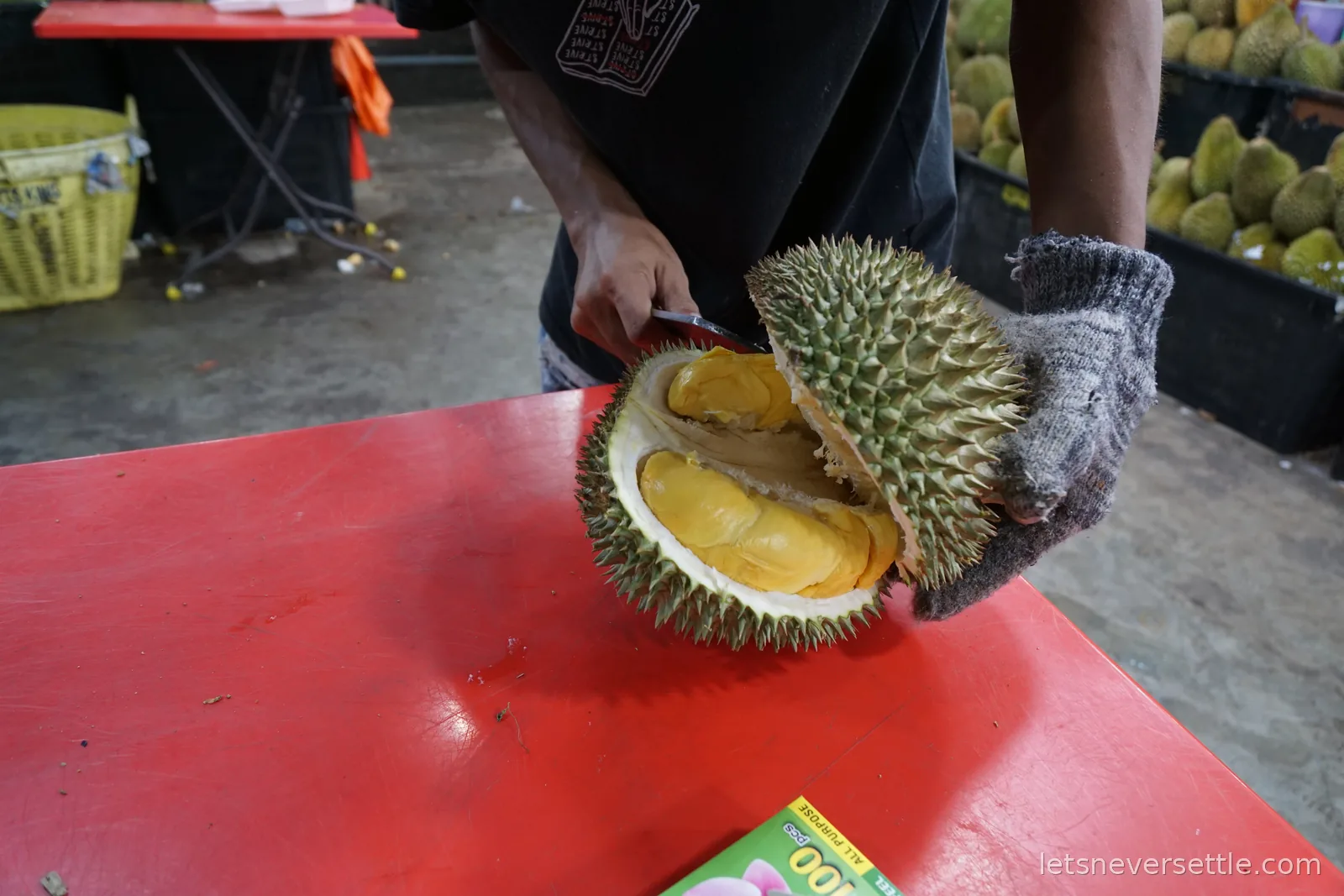 A vendor wearing a thick glove holding a durian open with a knife on a red table, showing the yellow flesh inside, with boxes of durians stacked behind