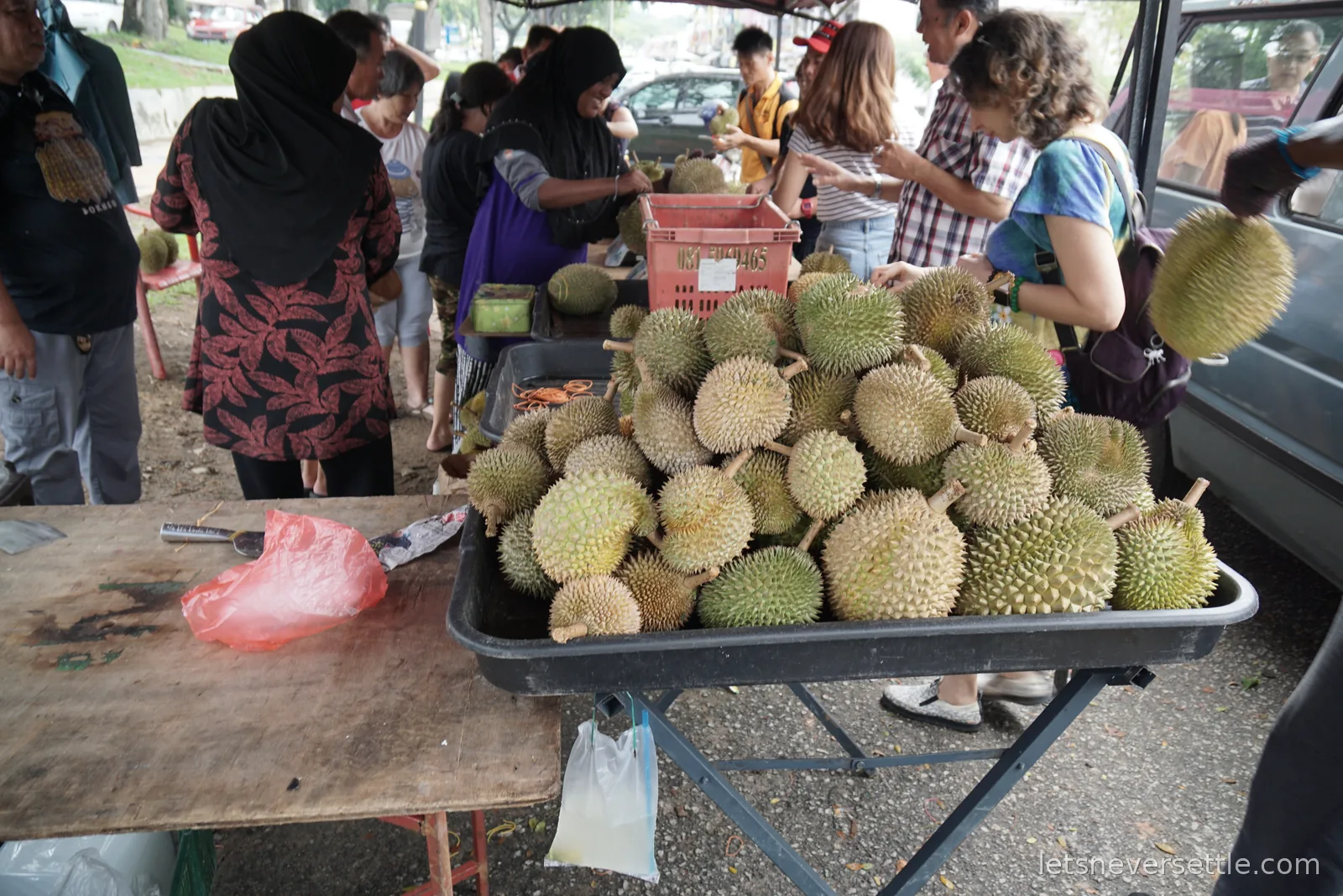 A roadside durian stall in Johor Bahru with a metal tray heaped with spiky durians, a crowd of customers around it and a vendor opening fruit at a table