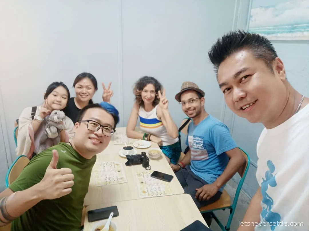 A group selfie of six people around a cafe table in Johor Bahru, several making peace signs, with a camera and menus on the table