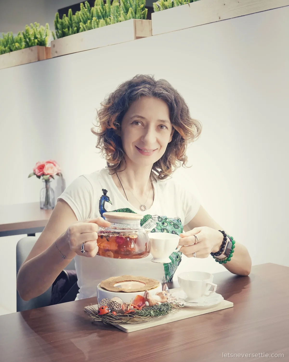 Inga sitting at a wooden cafe table in Johor Bahru pouring fruit tea from a glass teapot into a white cup, with a dessert on a board in front of her