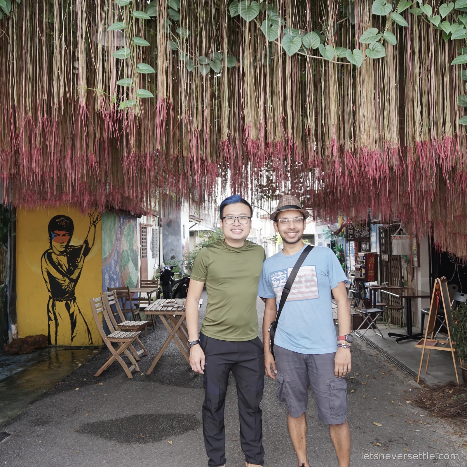 Dileep and a friend standing in a Johor Bahru shophouse lane under a hanging curtain of dyed pink and beige roots, with a Bruce Lee mural painted on the yellow wall behind