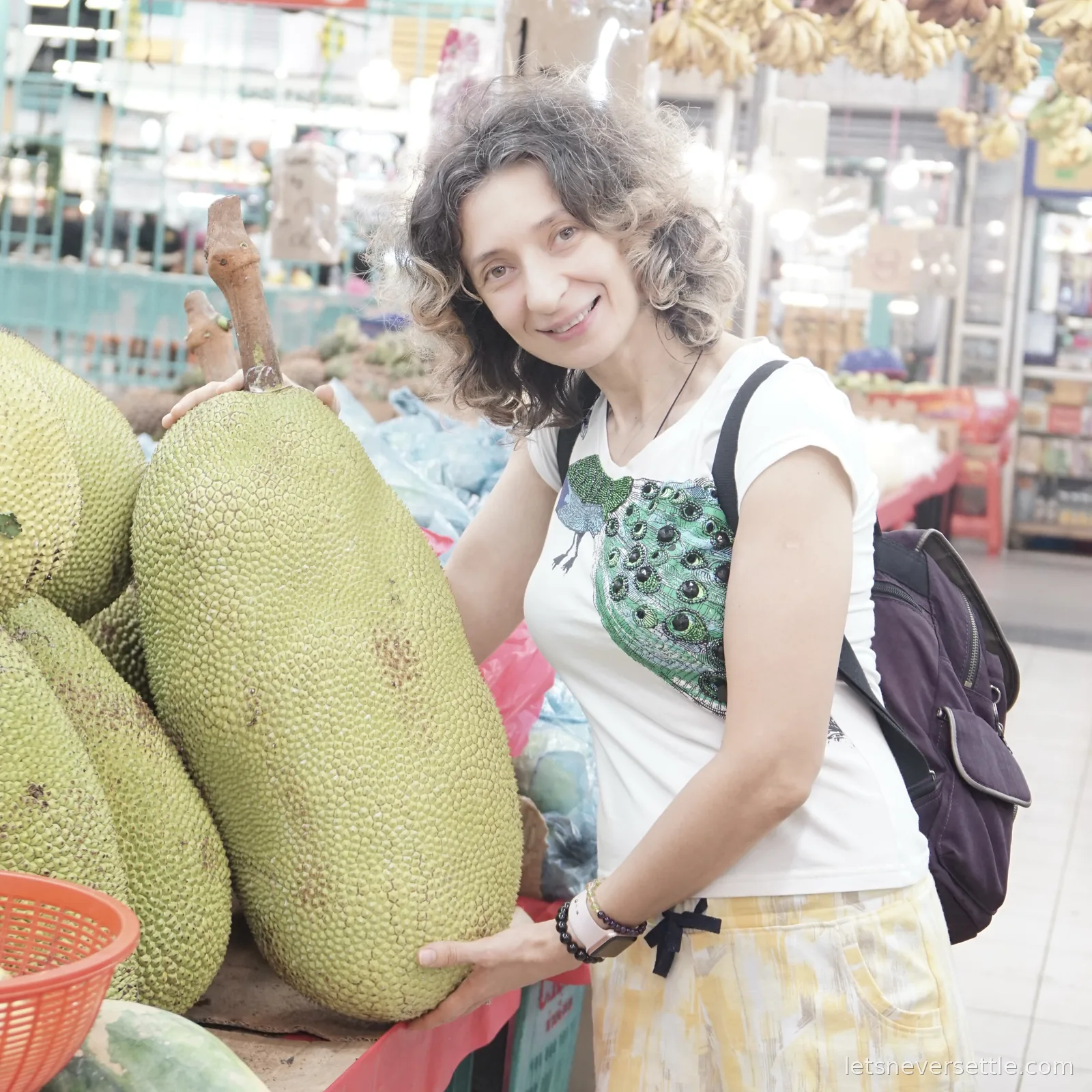 Inga holding up an enormous green jackfruit taller than her torso at an indoor market stall in Johor Bahru, with bunches of bananas hanging above