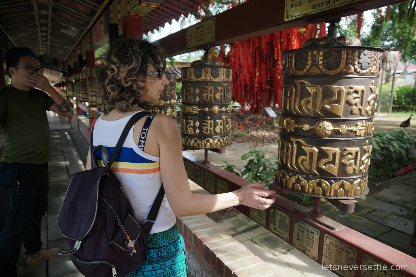 Inga turning a row of large brass prayer wheels under a covered temple walkway in Johor Bahru, with red prayer ribbons tied up behind them