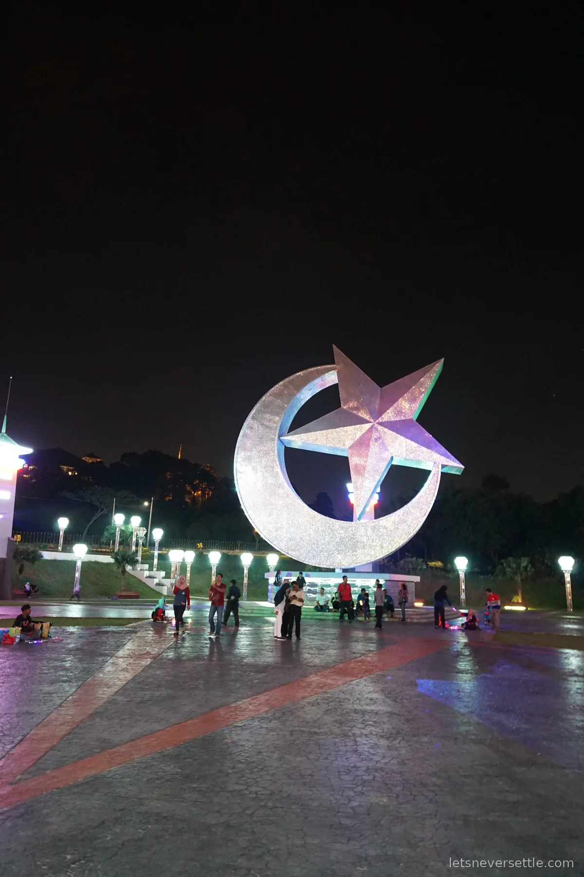 A large illuminated star and crescent sculpture standing in a public square in Johor Bahru at night, with people walking along a lit path behind it