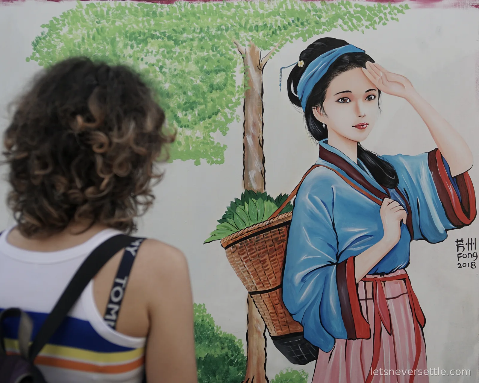 Inga seen from behind looking at a painted street mural of a woman in a blue jacket carrying a basket of greens on her back, signed Fong 2018