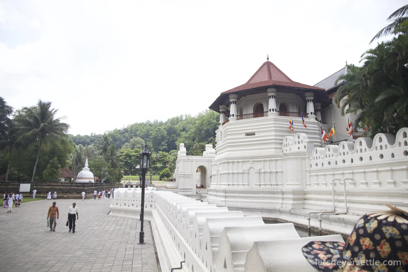White outer walls and octagonal red-roofed tower of the Temple of the Sacred Tooth Relic in Kandy, with a white stupa and palm trees behind