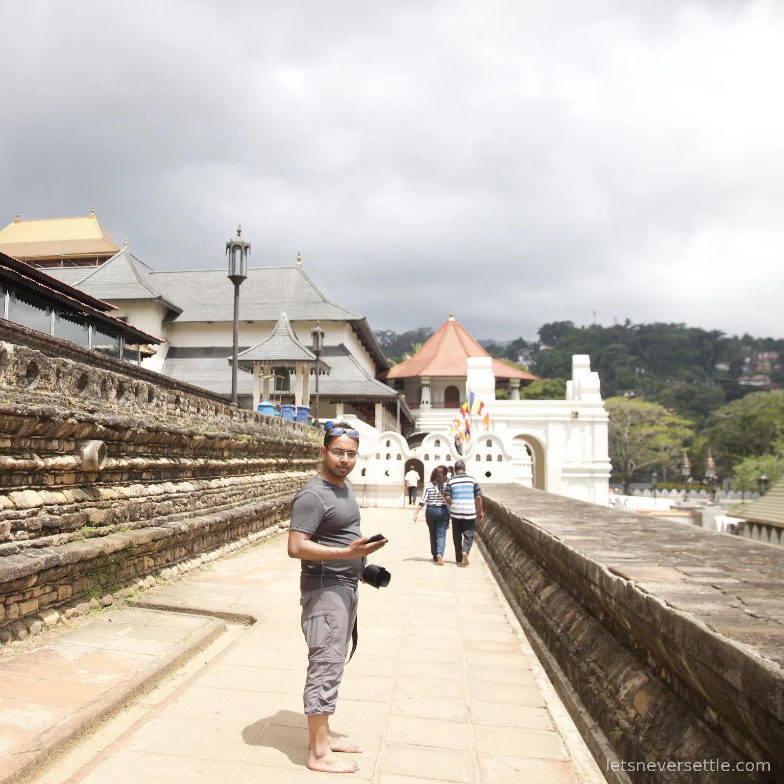 Dileep standing barefoot with a camera on the stone upper terrace beside the temple, a golden roof and hillside houses behind