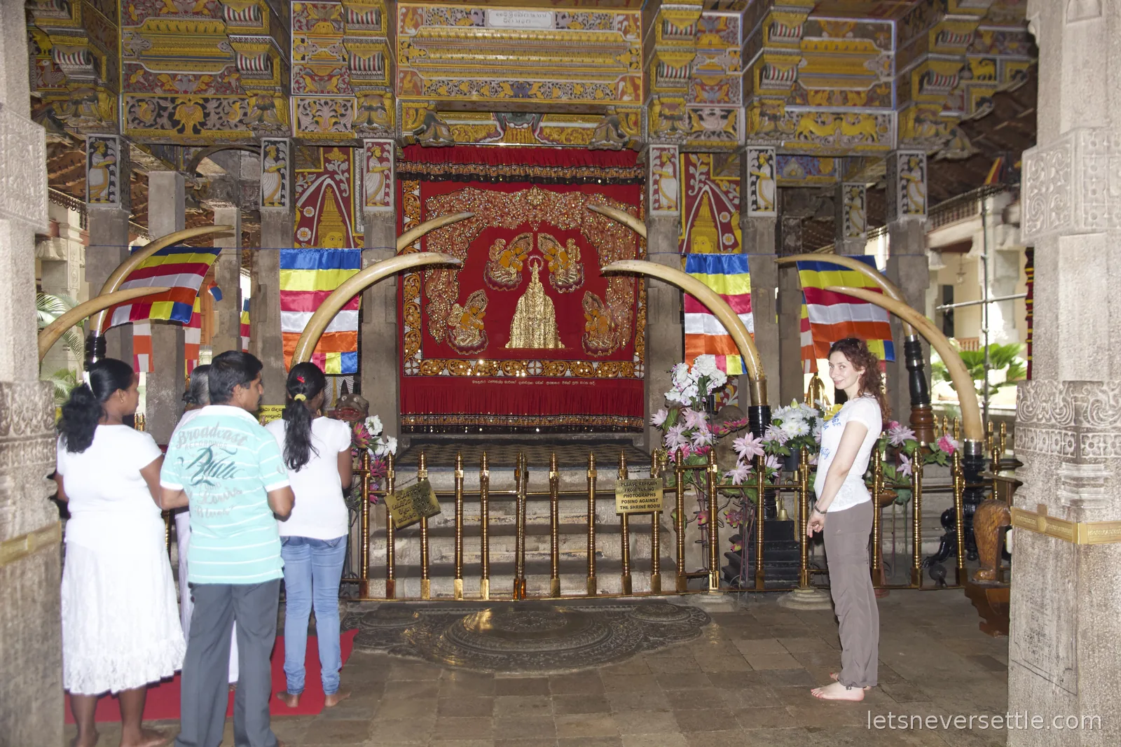 Worshippers standing before the red and gold shrine of the tooth relic, framed by curving elephant tusks and Buddhist flags