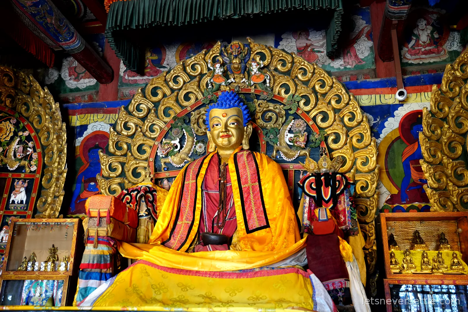 Gilded seated Buddha statue with blue hair and yellow silk robes inside a temple at Erdene Zuu, framed by an ornate golden halo and painted murals