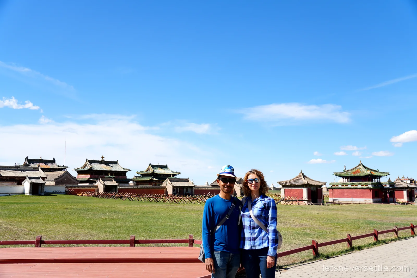 Dileep and Inga standing together on the grounds of Erdene Zuu monastery, Chinese-style temple roofs and white walls behind them under a big blue sky