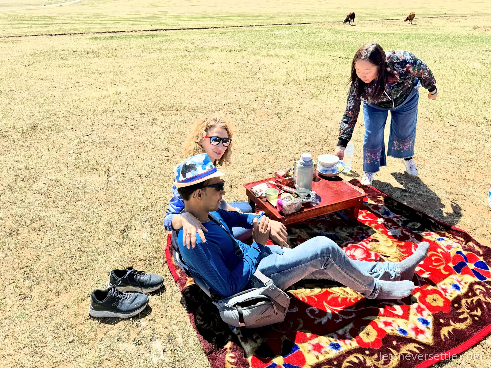 Dileep and Inga picnicking on a patterned blanket on the open steppe east of Kharkhorin, their guide setting out tea while cattle graze behind
