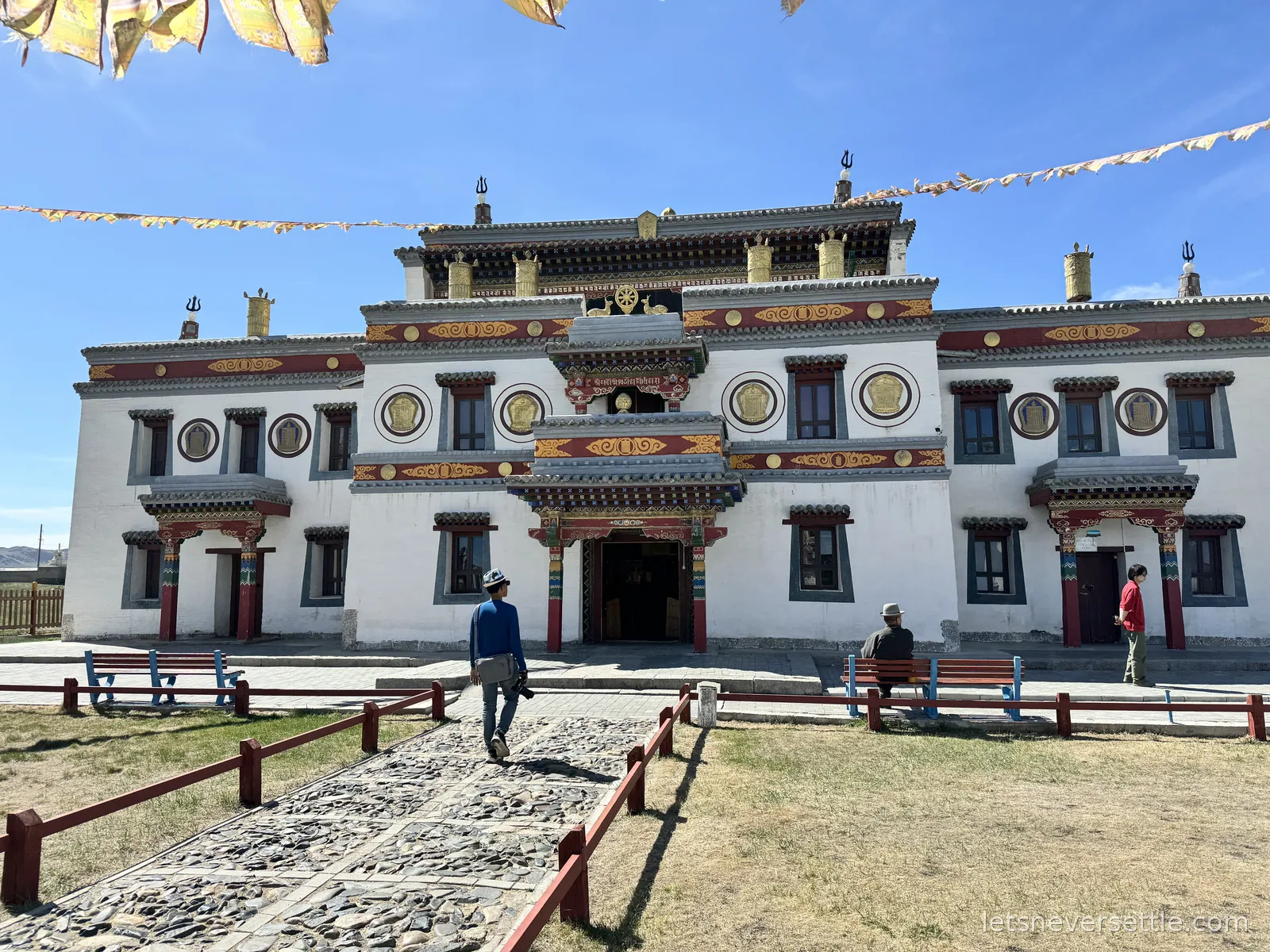 Dileep walking a stone path towards a white Tibetan-style temple at Erdene Zuu, strings of prayer flags overhead and gilded ornaments on the roofline