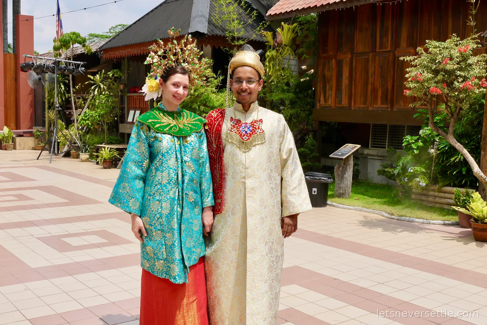Inga and Dileep dressed in traditional Malay royal wedding costumes in front of wooden kampung houses