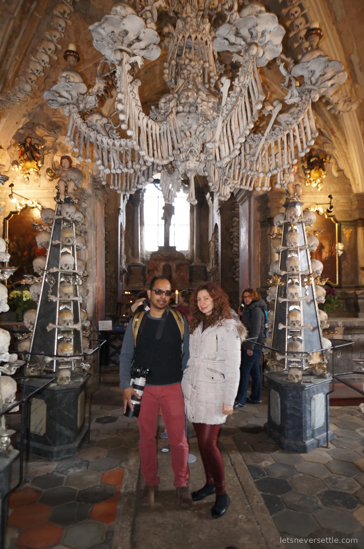 Inga and Dileep standing inside the Sedlec Ossuary between pillars topped with skulls and bones, a bright window behind them