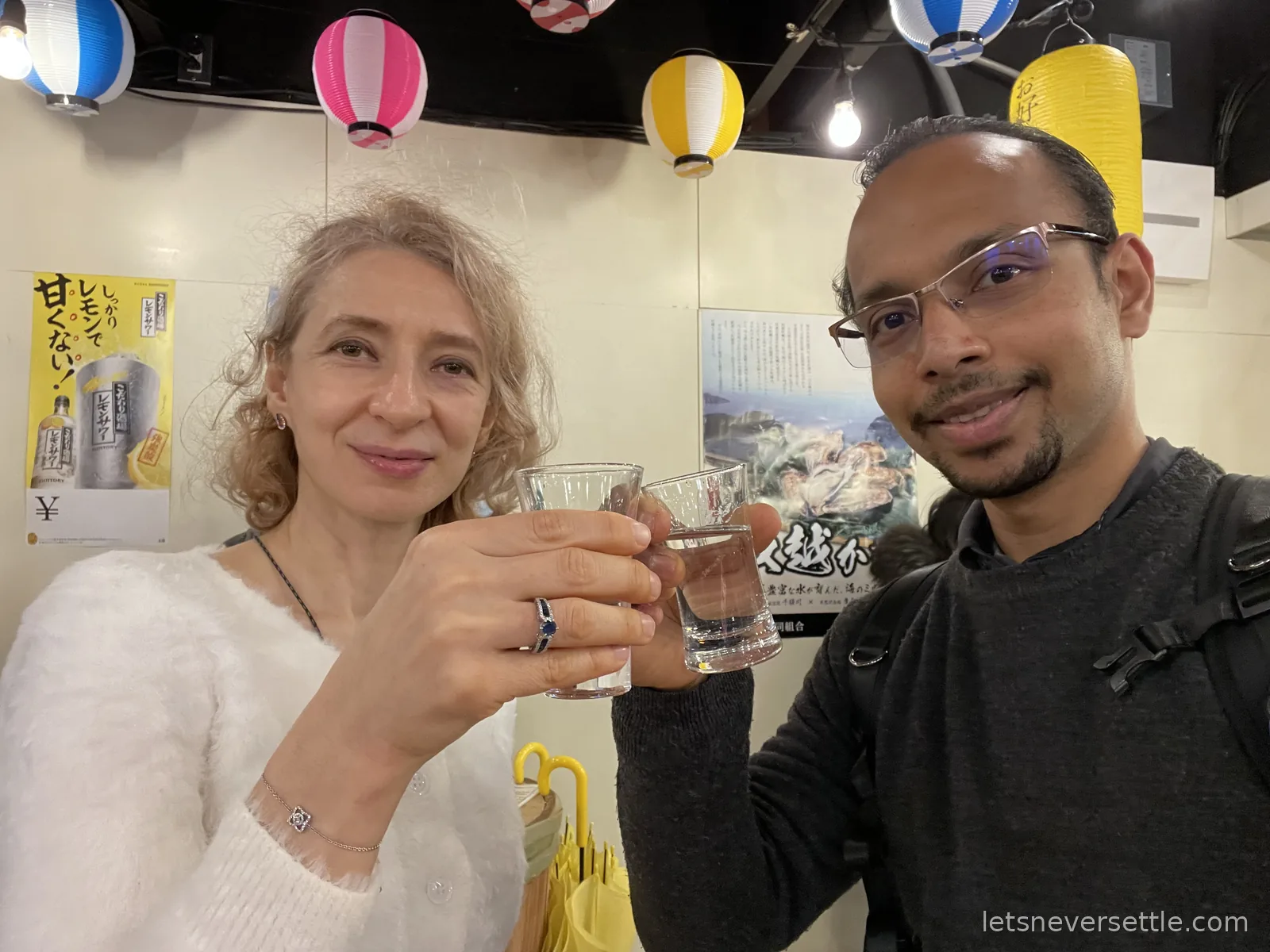 Inga and Dileep raising small glasses of sake under colourful paper lanterns at a market tasting counter