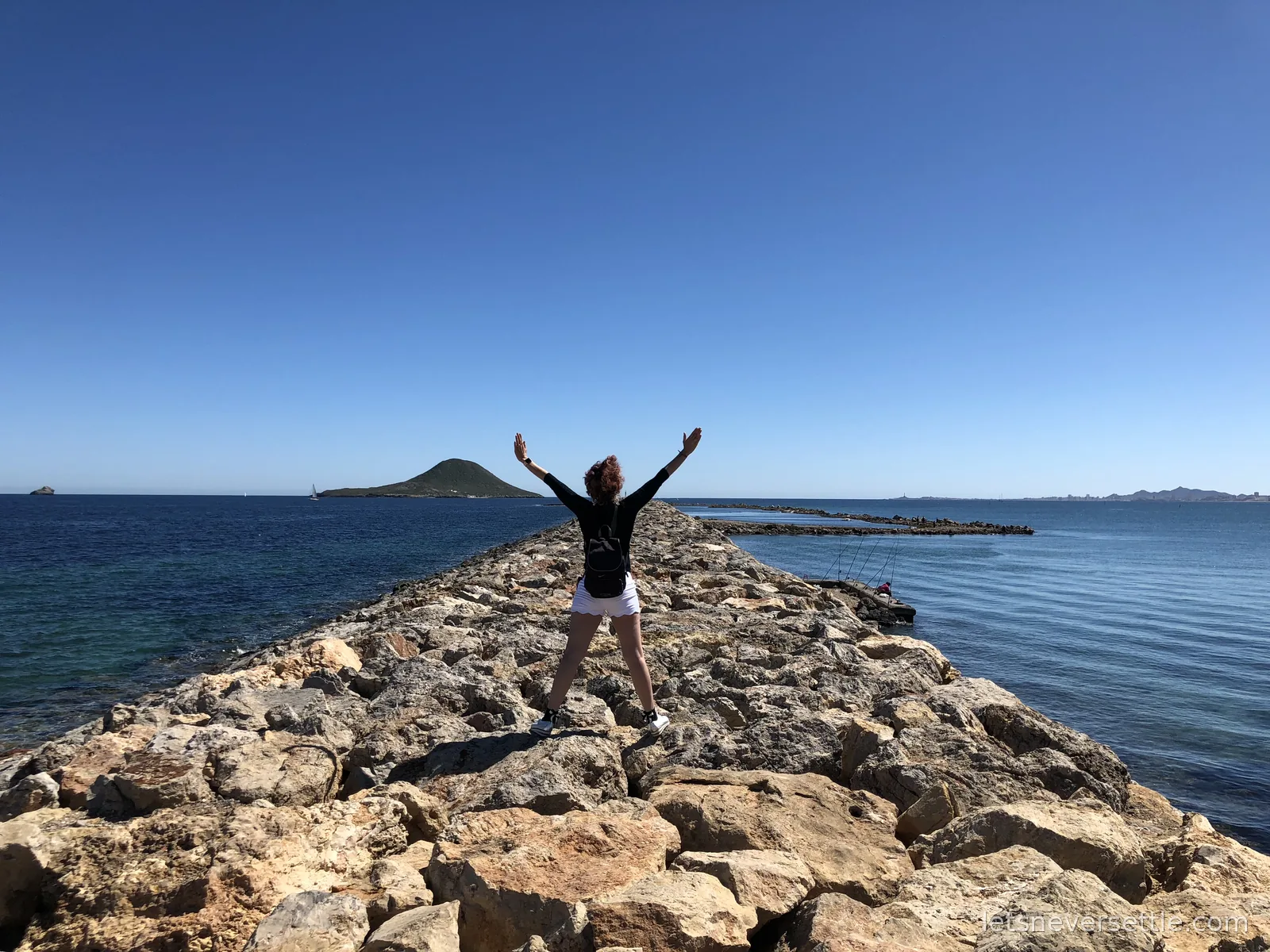 Inga with arms spread wide on a rocky breakwater stretching into calm blue sea, a conical island on the horizon