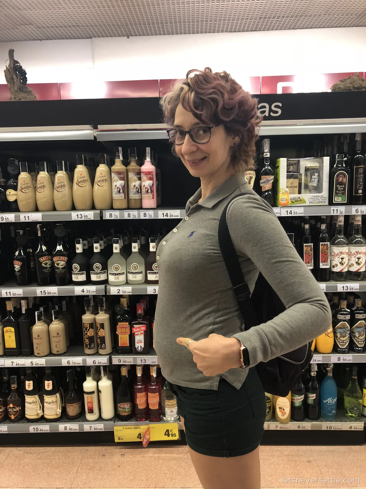 Inga gesturing with a thumb at supermarket shelves stacked with Spanish cream liqueurs and spirits in Cartagena