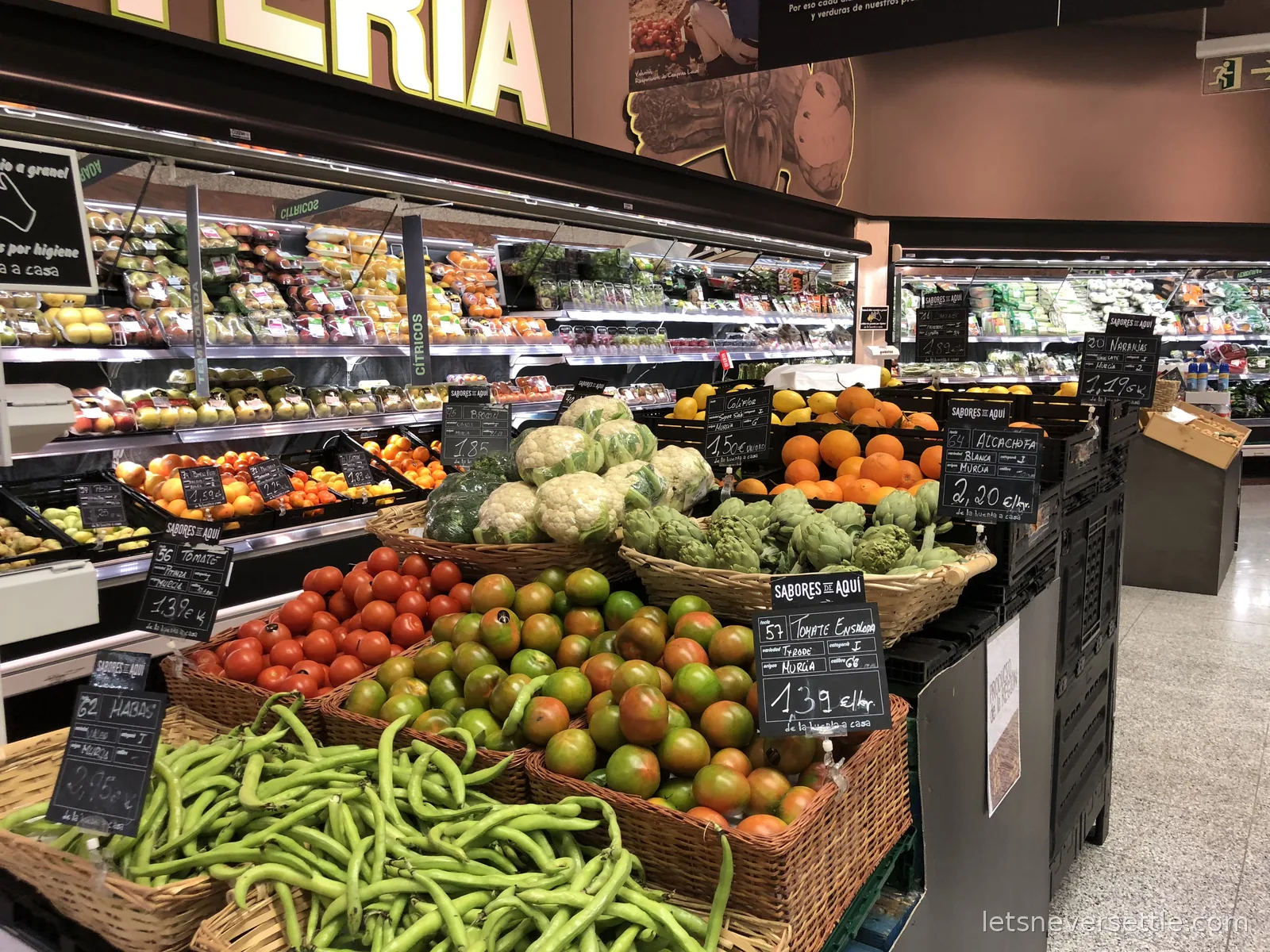 Baskets of broad beans, tomatoes and artichokes with handwritten chalkboard labels naming Murcia as origin in a supermarket