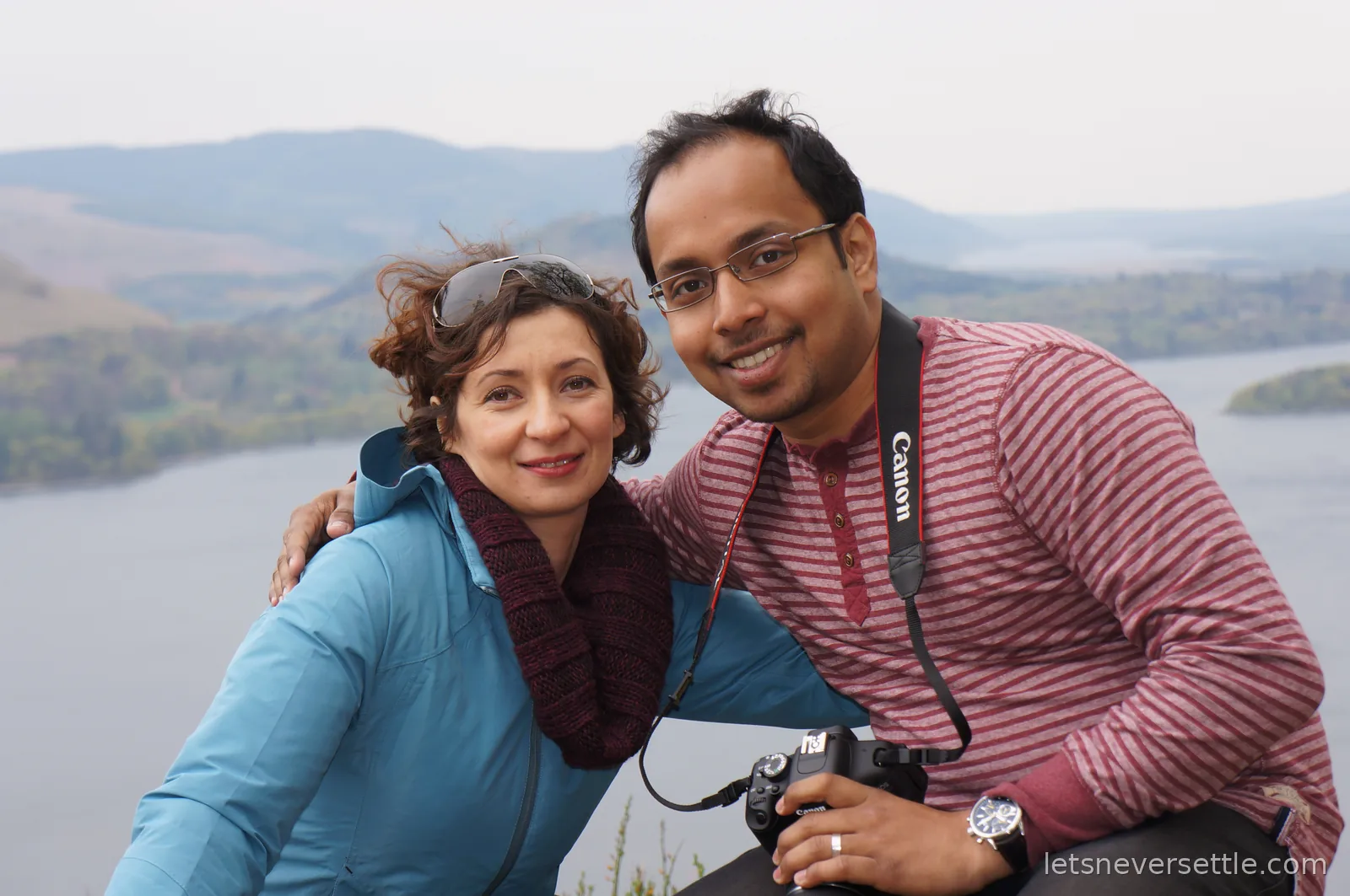 Inga and Dileep close together high above a broad lake, wooded shores and misty fells stretching into the distance