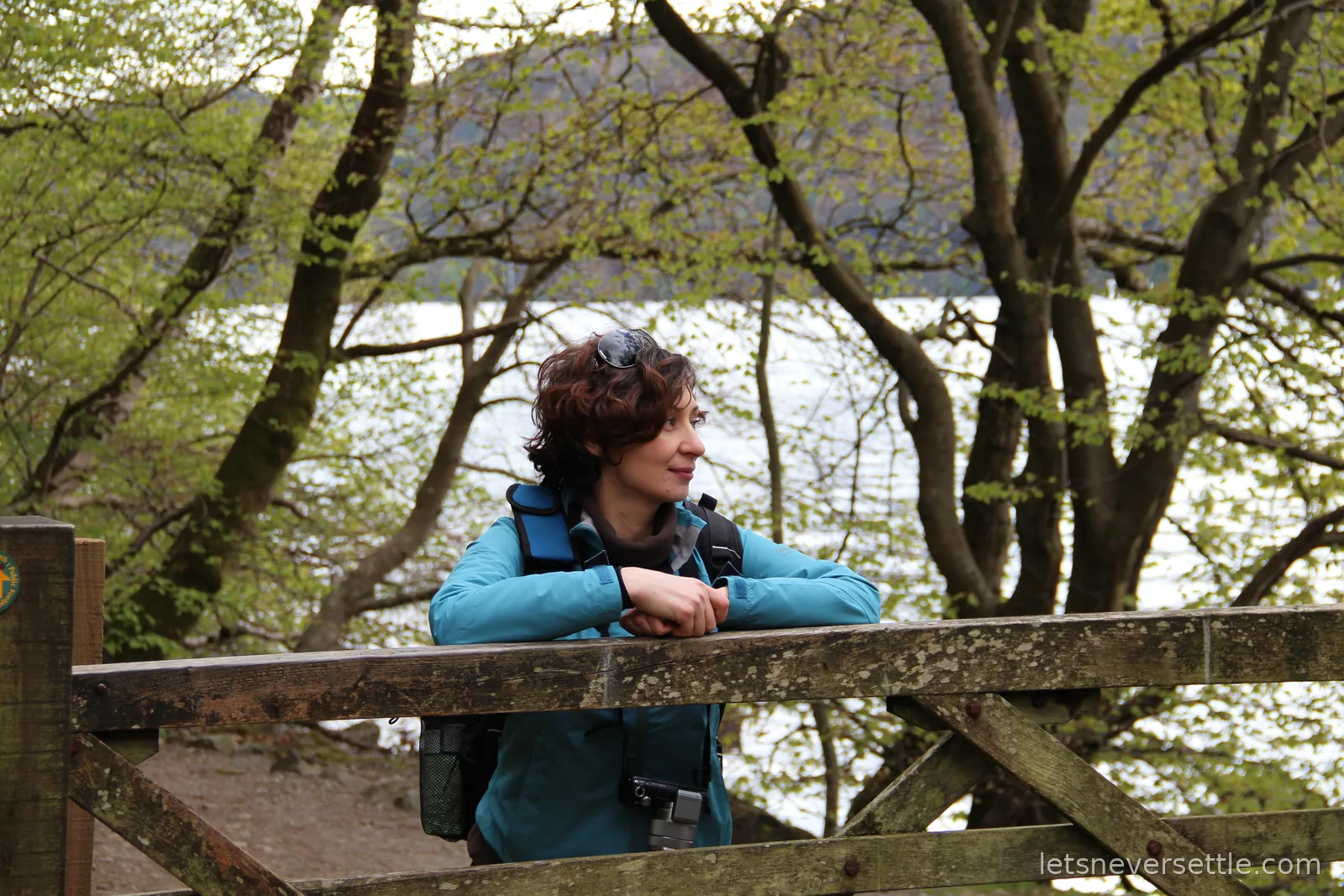 Inga leaning on a wooden gate under trees at the edge of the lake, the water bright behind her