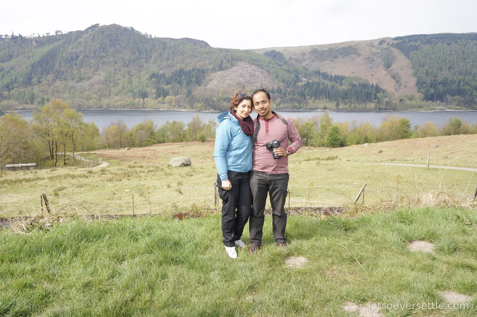 Inga and Dileep standing together in a lakeside field, forested fells rising across the water behind them