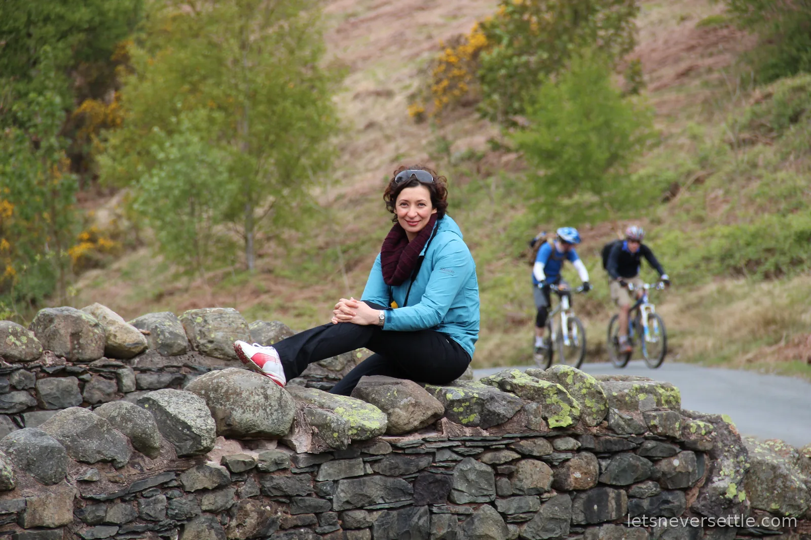 Inga sitting on a mossy stone wall by a fellside lane while two mountain bikers ride past behind her