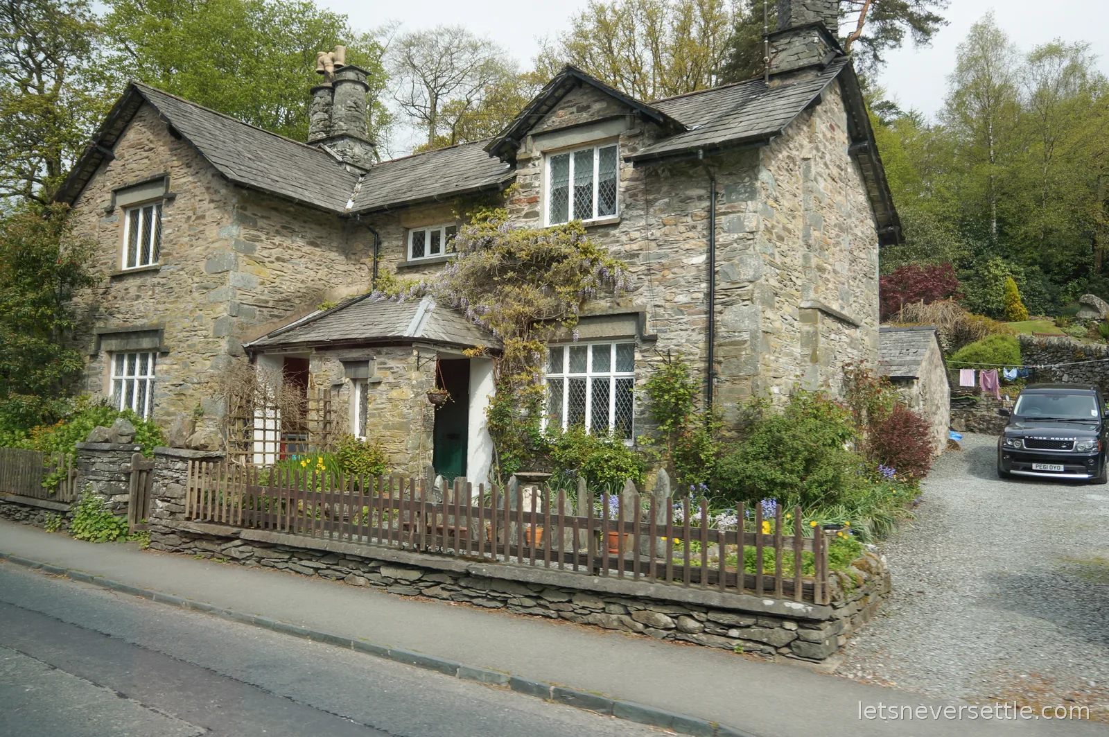 A Lakeland stone cottage with wisteria climbing over the porch, spring flowers along the fence and a gravel drive beside it