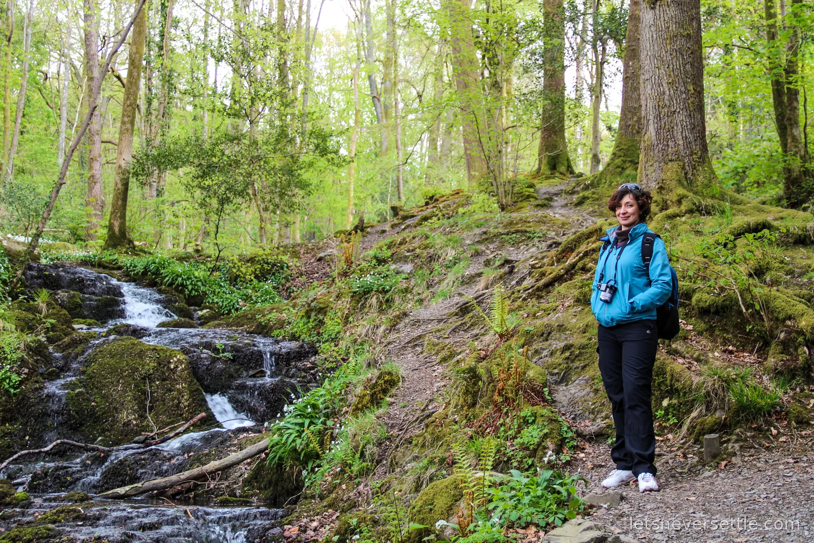 Inga in a blue jacket on a woodland path beside a small waterfall tumbling over mossy rocks between spring green trees