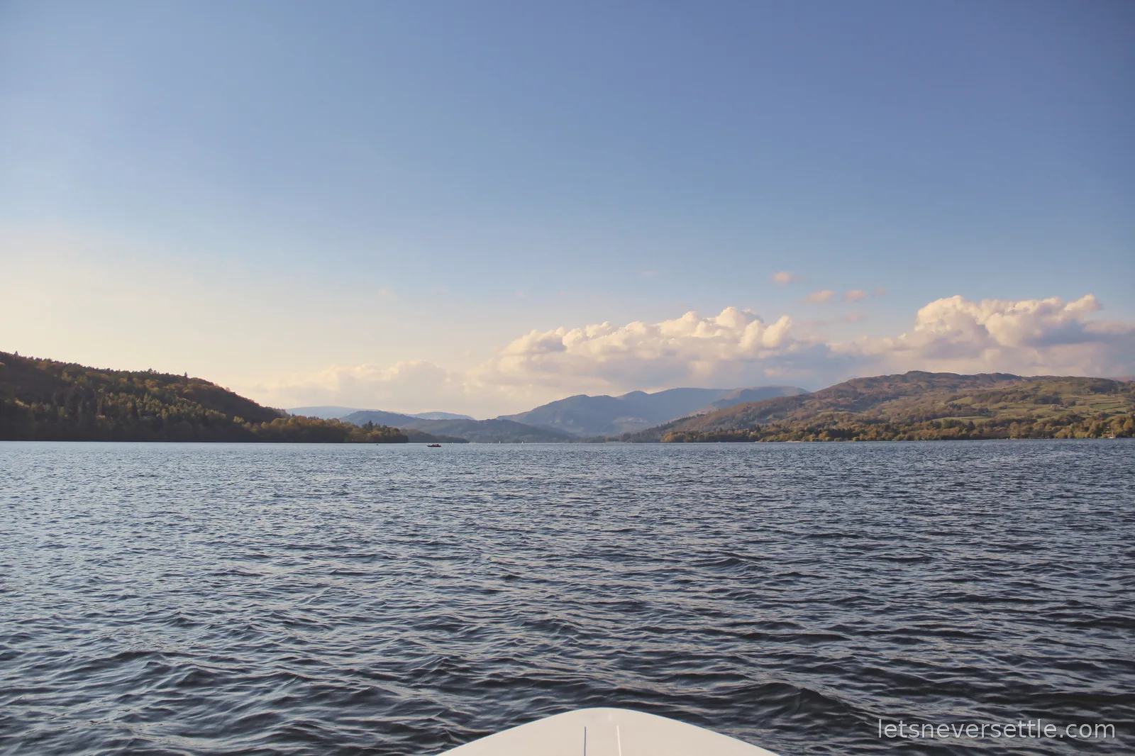 View over the bow of a small boat down the length of Windermere, fells rising along both shores under a blue evening sky