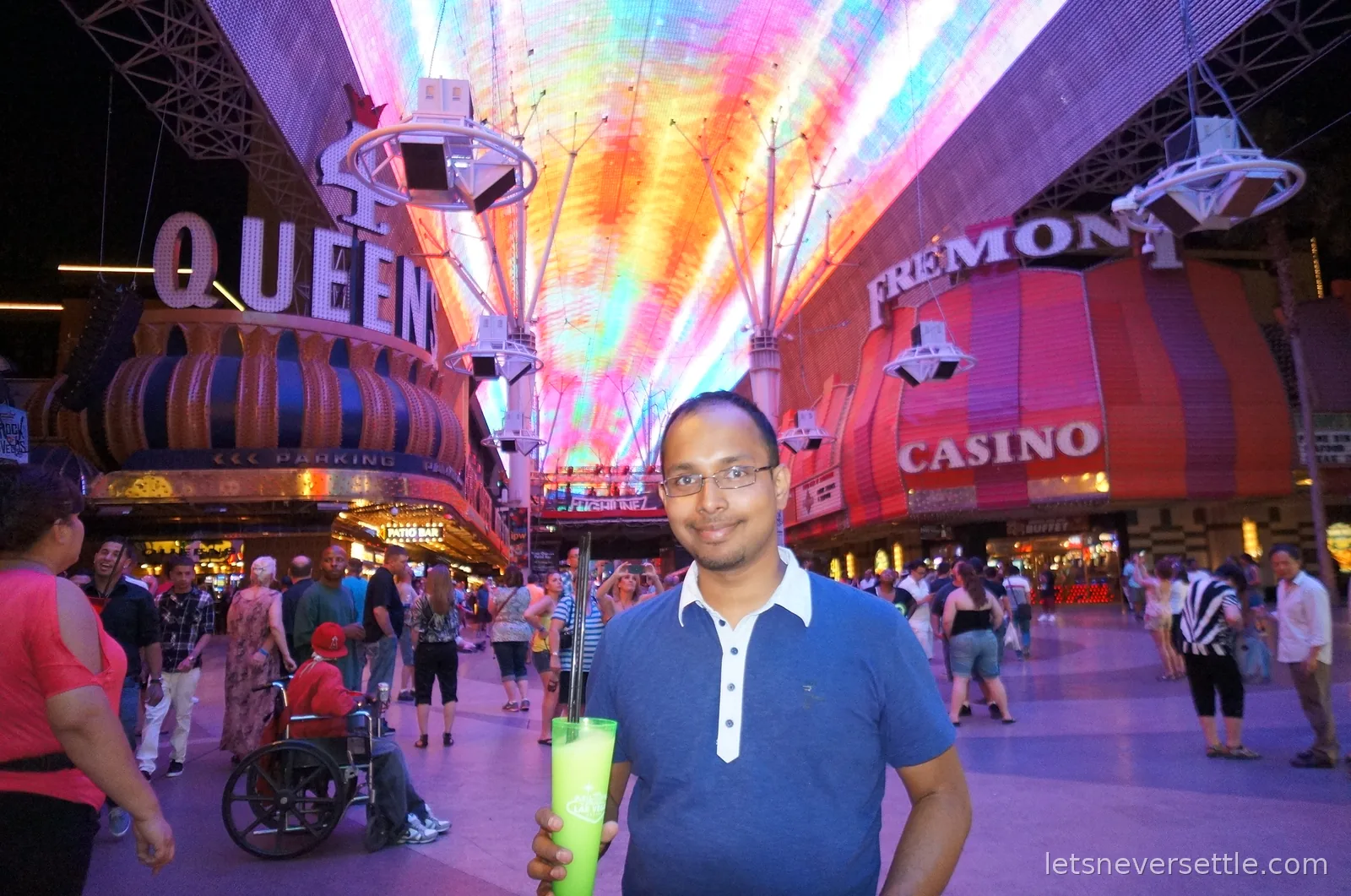 Dileep holding a green souvenir drink under the glowing LED canopy of Fremont Street, Four Queens and Fremont casino signs either side