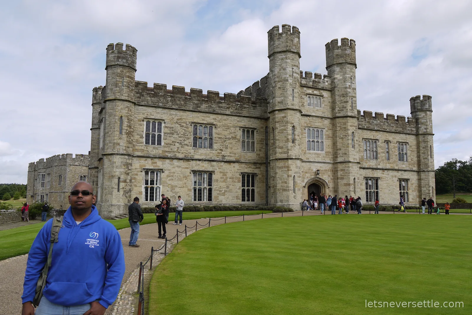 Dileep's brother in a blue university badminton club hoodie in front of the battlemented main front and lawns of Leeds Castle