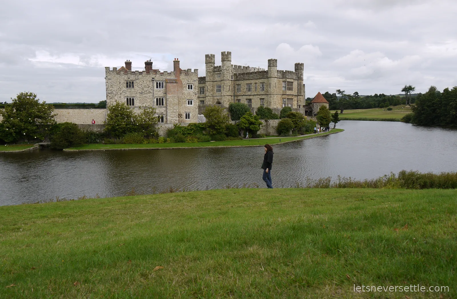 Leeds Castle seen across its moat under a grey sky, with Inga walking along the grassy bank in the foreground