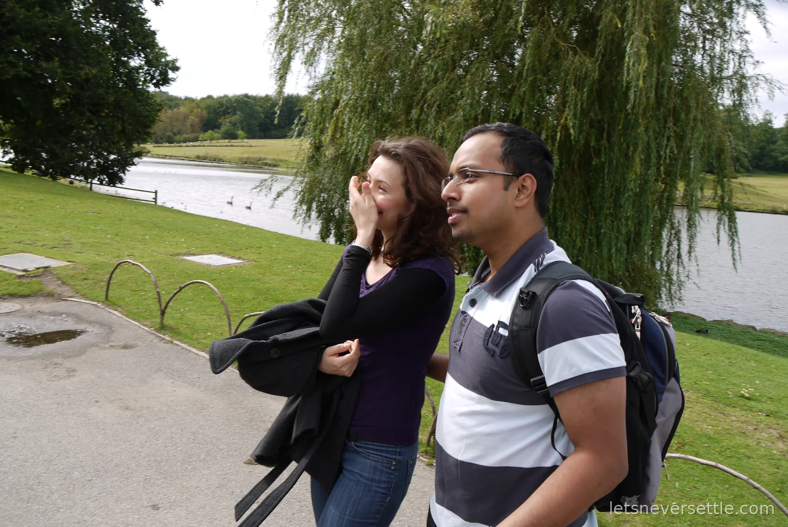 Inga covering her mouth and laughing as she walks with Dileep along the moatside path under a willow at Leeds Castle