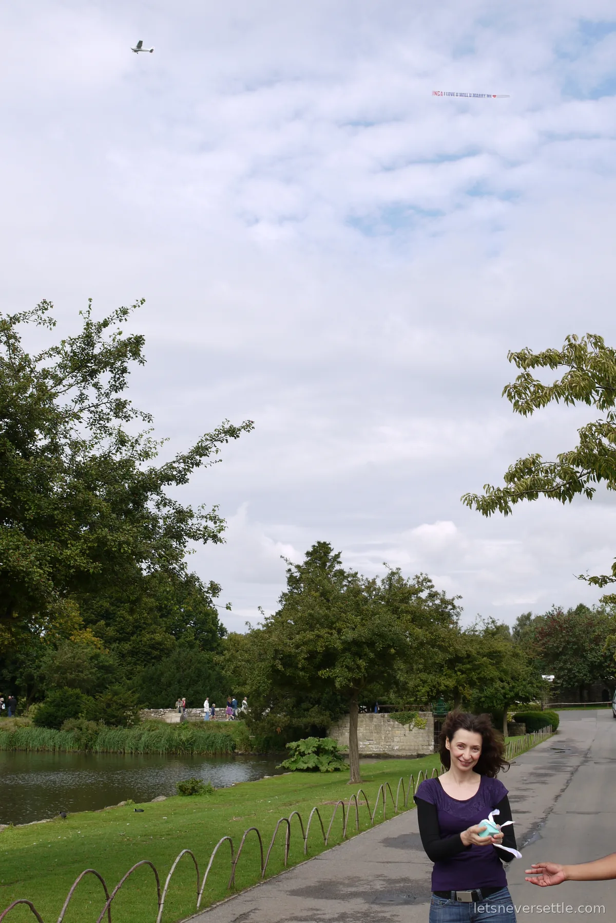 Inga smiling on the path by the moat holding a small ring box, while a light aircraft tows a marriage proposal banner high above the trees