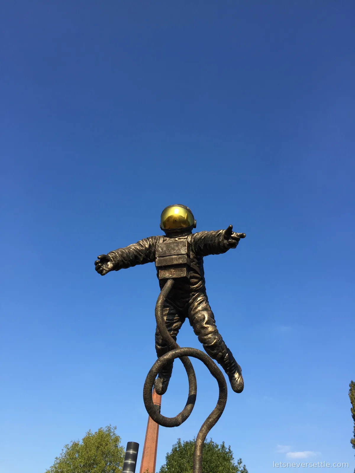 A bronze astronaut statue with a gold mirrored visor and a coiled umbilical line, photographed against a clear blue sky