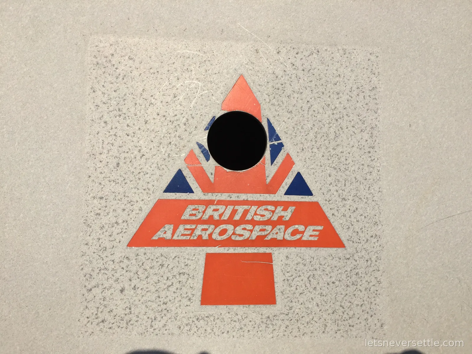 A worn British Aerospace sticker with a red and blue arrowhead logo on pale speckled spacecraft panelling