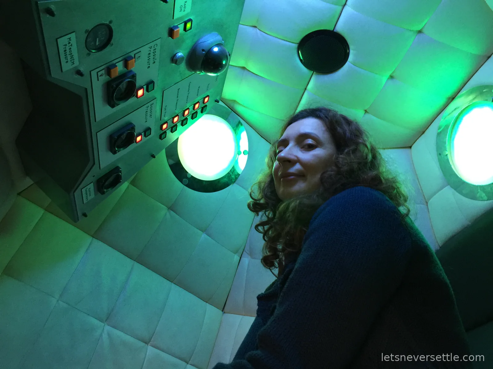 Inga inside the padded capsule mock-up, lit green through the portholes, beside a control panel labelled Capsule Pressure and Booster Pressure