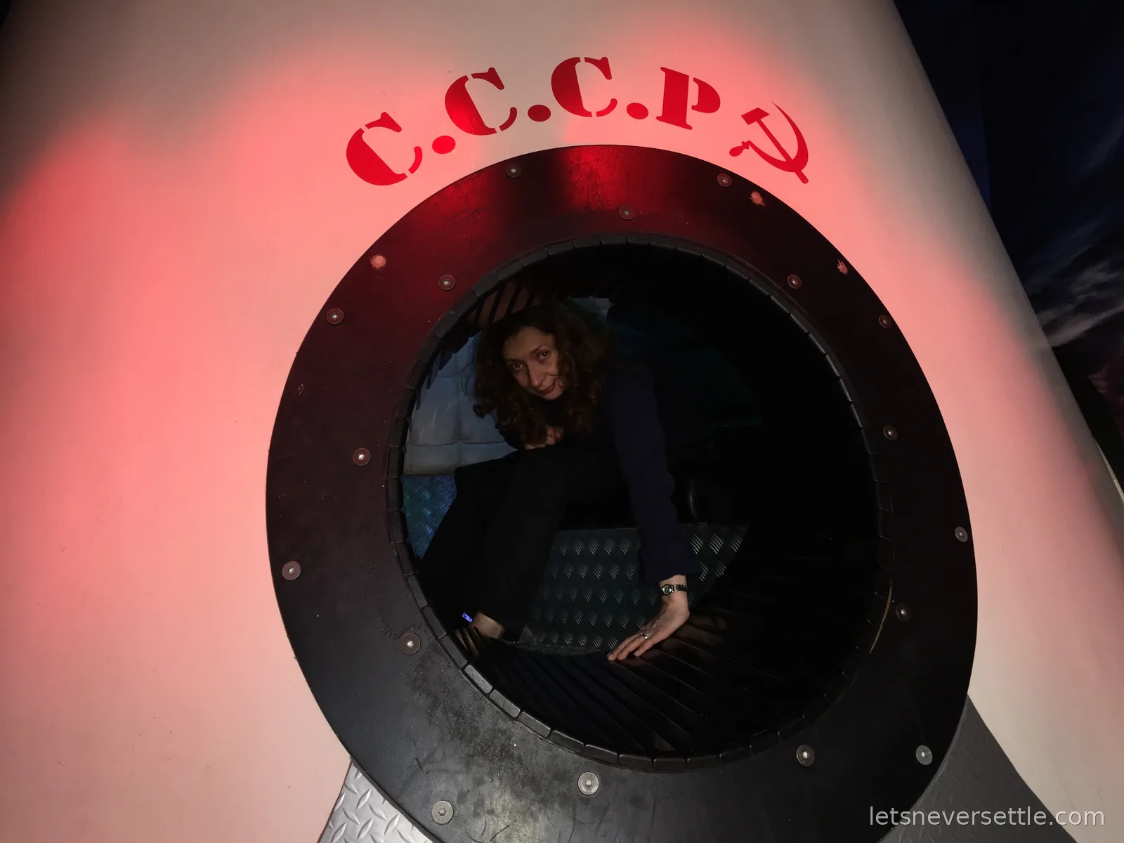 Inga crouched inside the round hatch of a white space capsule mock-up stencilled C.C.C.P with a red hammer and sickle
