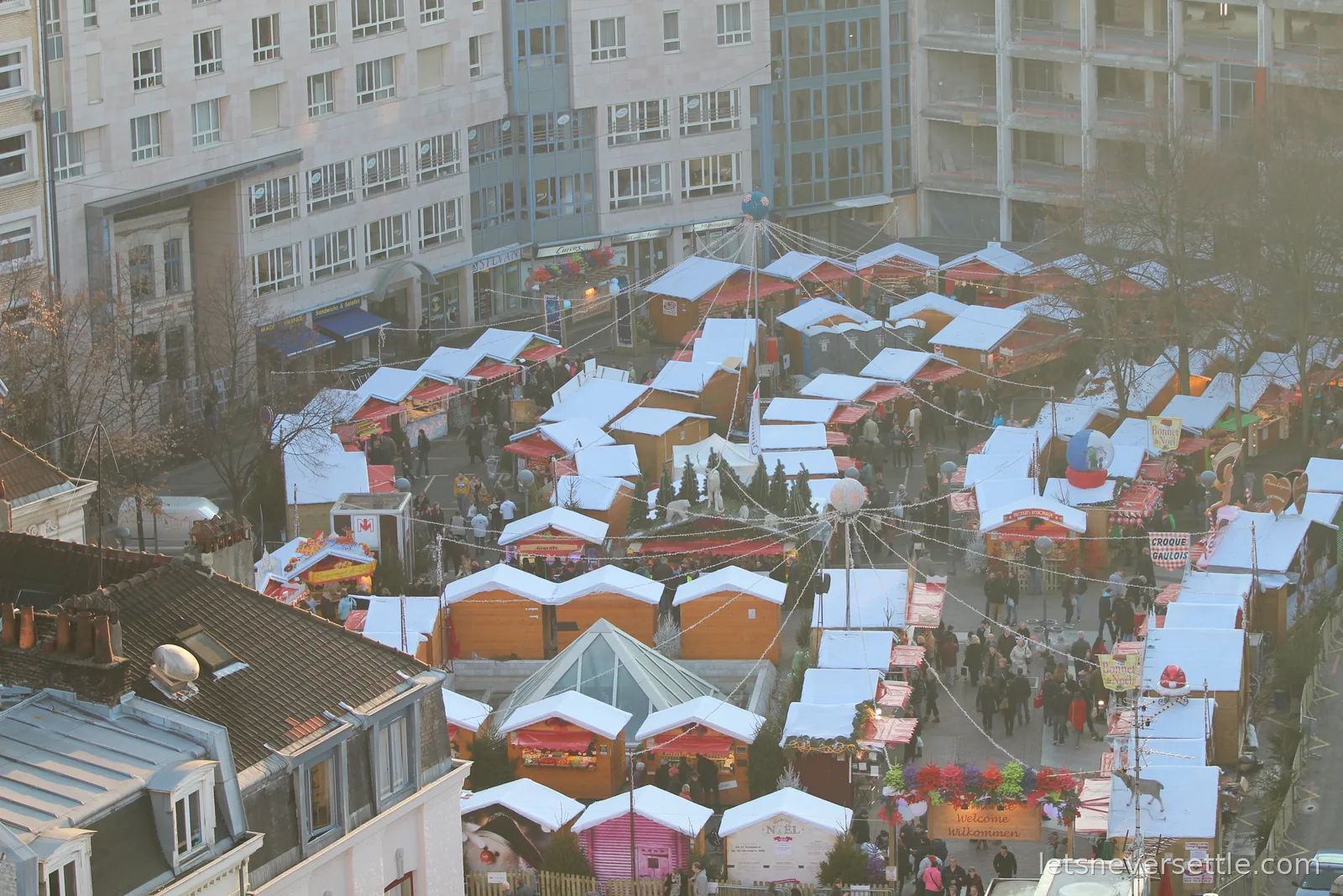 Aerial view of the Lille Christmas market, rows of wooden chalets with white roofs and strings of lights filling a square between buildings
