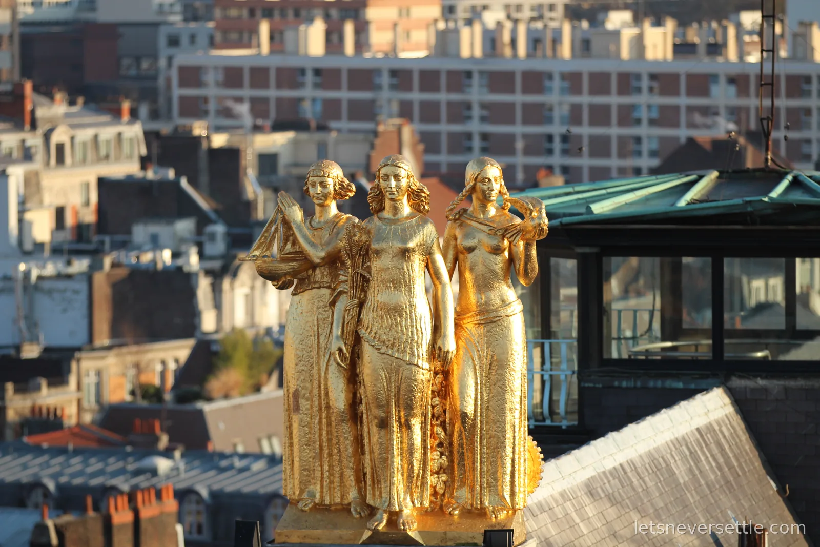 Three gilded female statues on a Lille rooftop catching the evening sun, one holding a sailing ship, city buildings soft behind