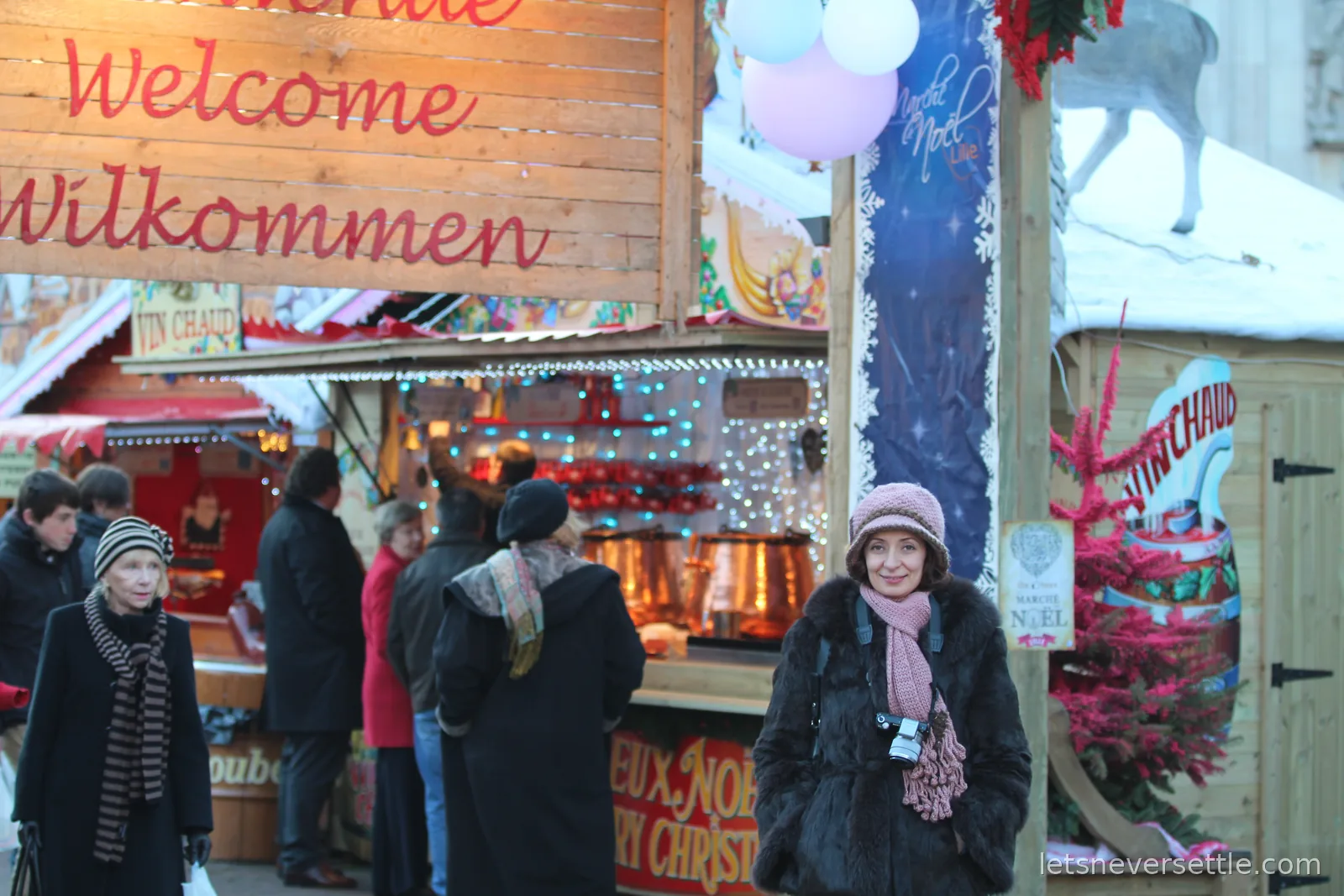Inga in a pink knitted hat and fur coat at the entrance of the Lille Christmas market, welcome signs and copper pots of vin chaud behind