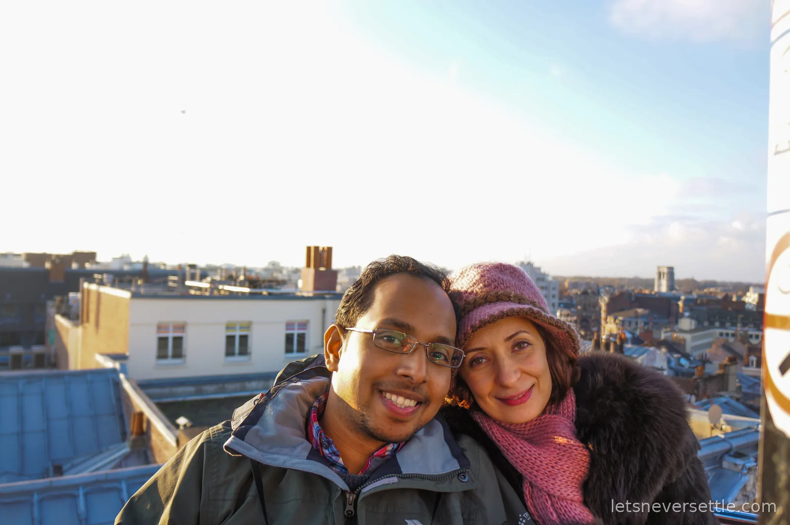 Inga in a pink knitted hat and Dileep in glasses posing high above the rooftops of Lille, low sun flaring behind them