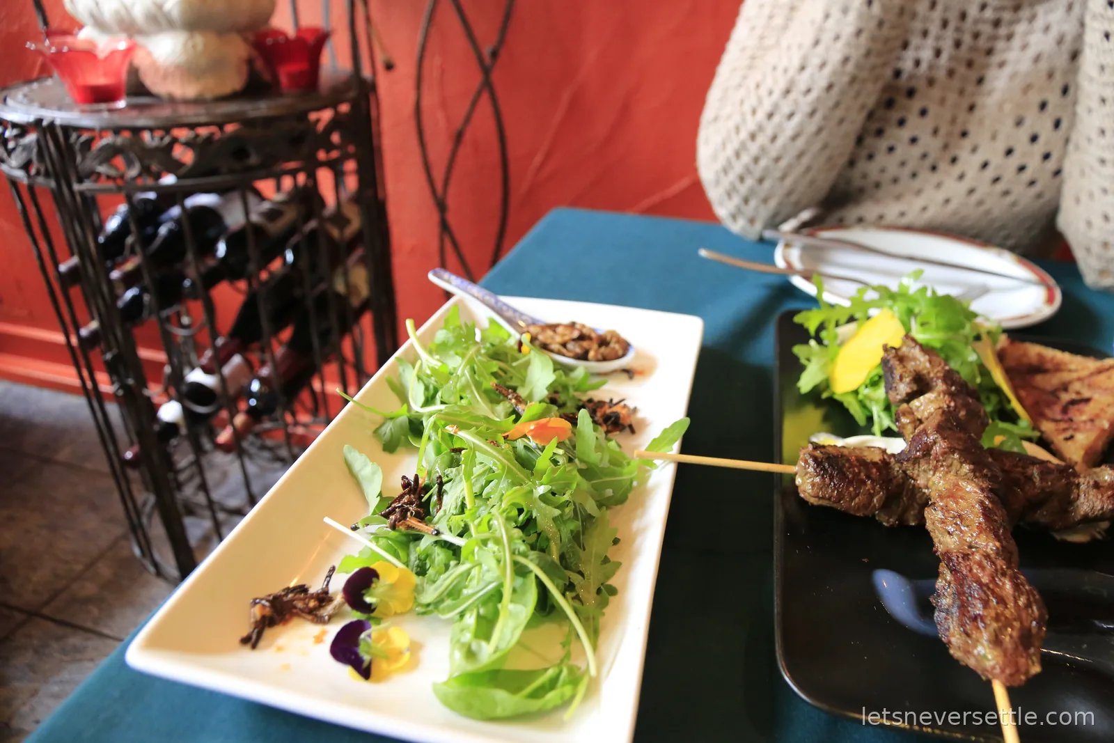 Two plates on a turquoise tablecloth, a long dish of green salad scattered with whole insects and a board of grilled skewers