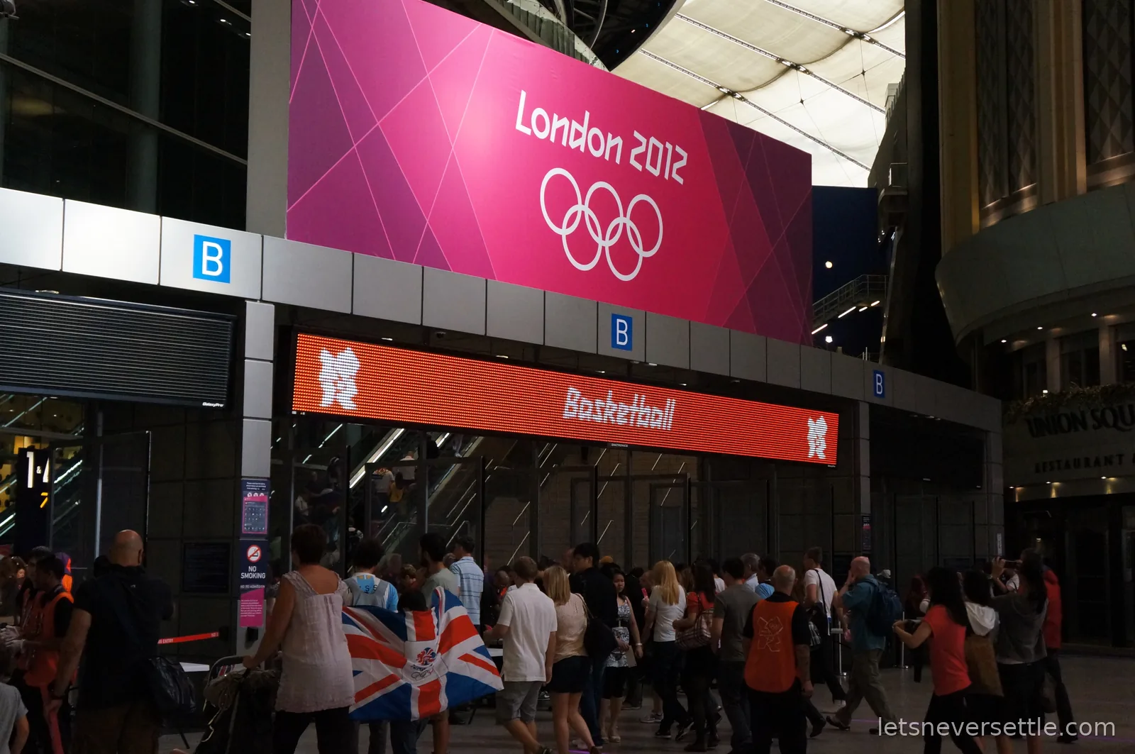 Crowds beneath a pink London 2012 banner and an orange Basketball sign at the arena entrance, a Union Jack draped over one visitor