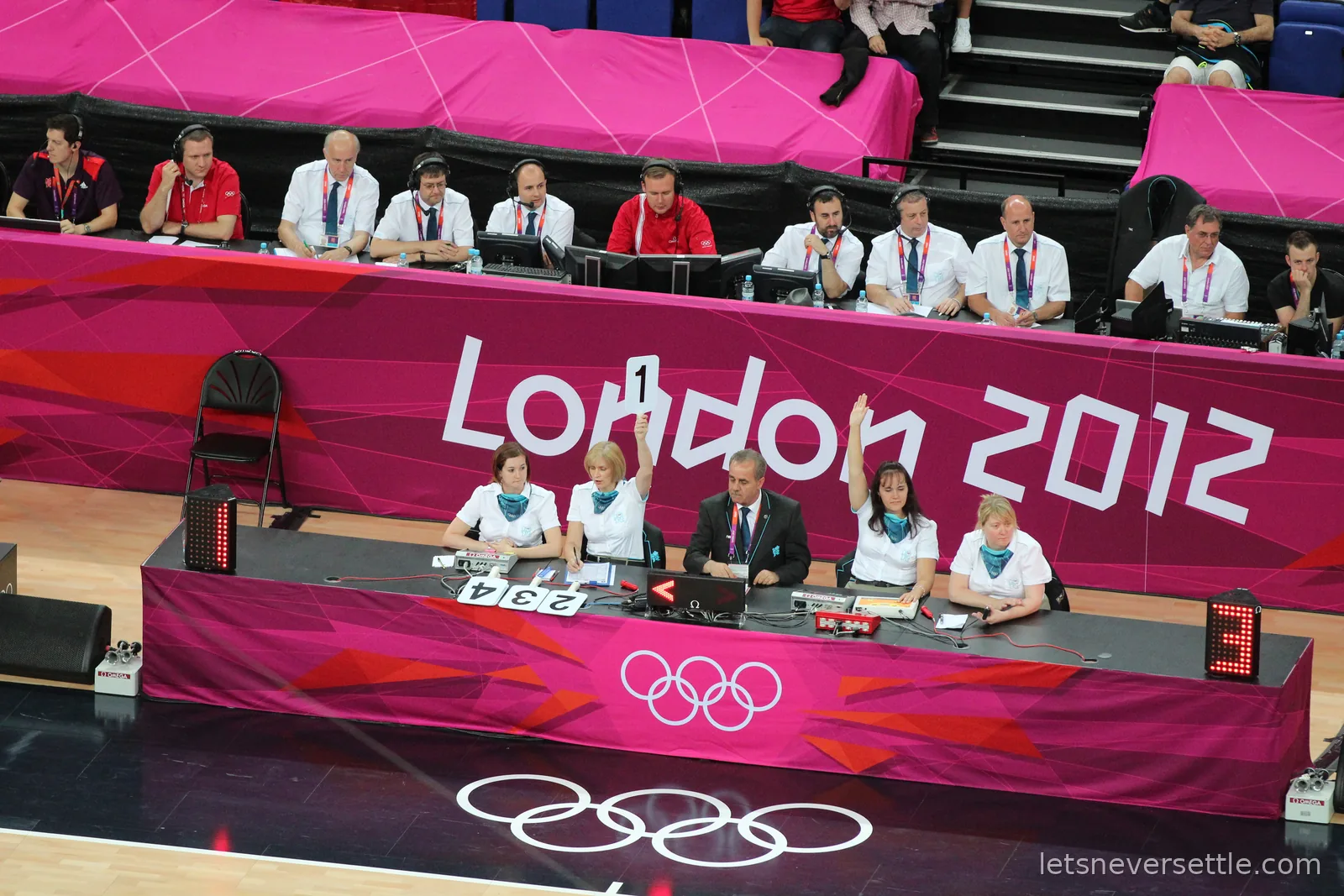 The scorers' table at the London 2012 basketball, officials with headsets in front of a pink London 2012 branded court